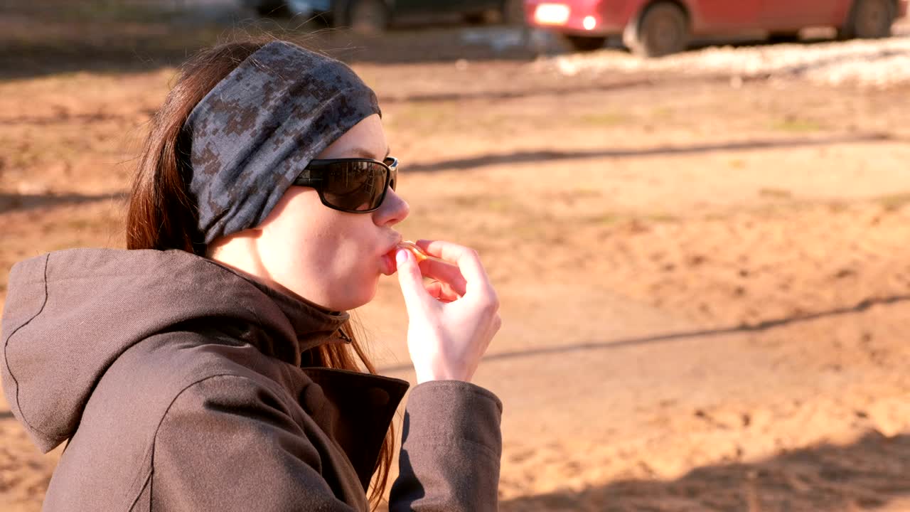 선글라스를 입은 젊은 갈색 여성이 공원의 벤치에 앉아 망가린을 먹습니다. 측면 시각.