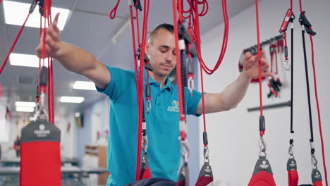 personal trainer controls the process of recovery client on suspension ropes. young man is hanging on special rope system in rehabilitation center. instructor helps his patient to get better.