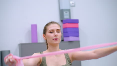 una mujer enfocada hace ejercicio estirando una banda de resistencia con los brazos levantados en un gimnasio bien iluminado, demostrando entrenamiento de fuerza y flexibilidad