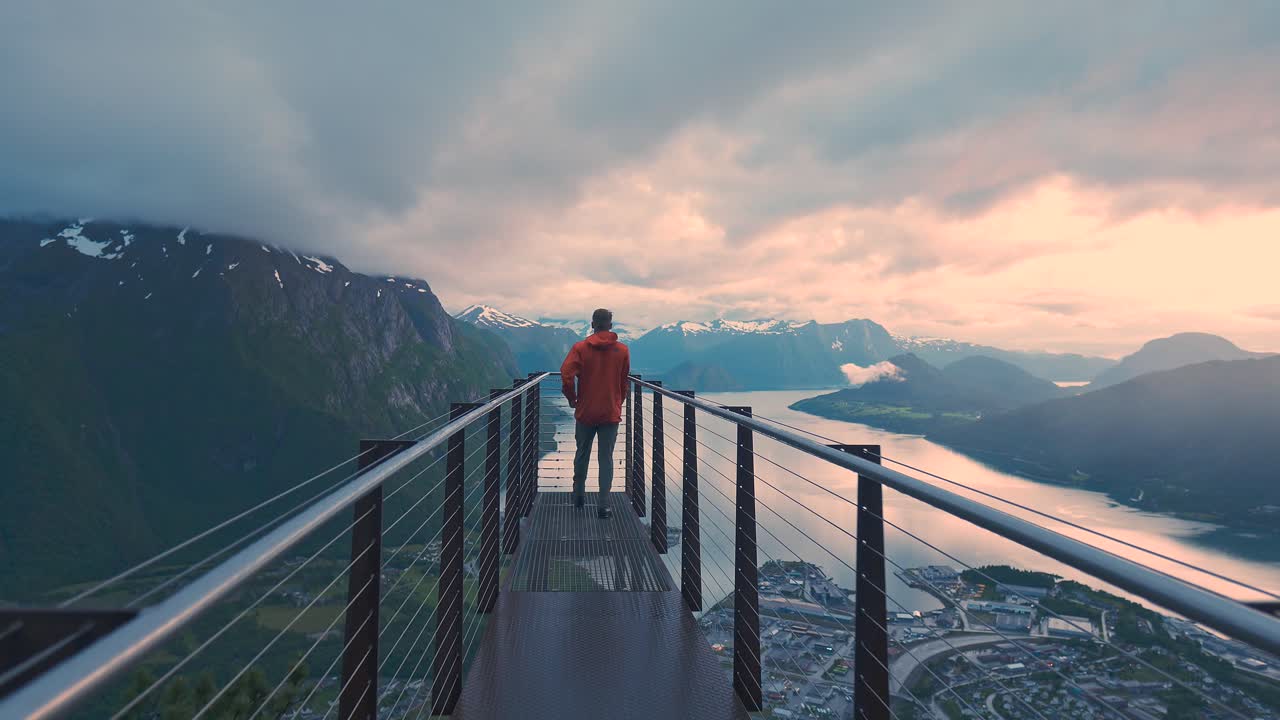 Man walking towards a beautiful view point, revealing incredible scenery during sunset. Watching over glorious mountain and sea view, next to a city with orange clouds in the sky. Norway Rampestreken.
