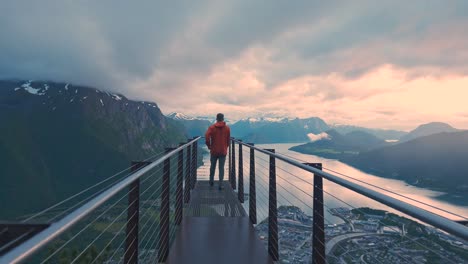 man walking towards a beautiful view point, revealing incredible scenery during sunset. watching over glorious mountain and sea view, next to a city with orange clouds in the sky. norway rampestreken.