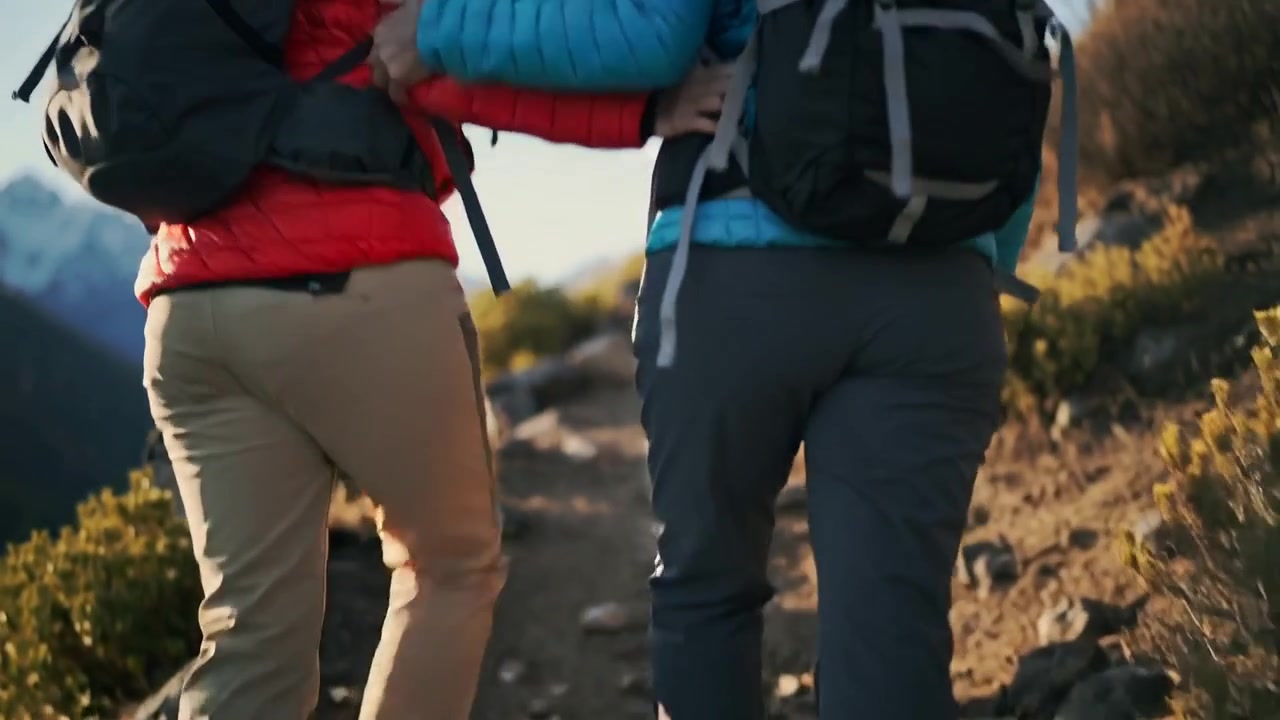 Una coppia di escursionisti percorre un sentiero alpino; quando la pendenza si fa più ripida, si tengono per braccio per darsi stabilità, con zaini e giacche.