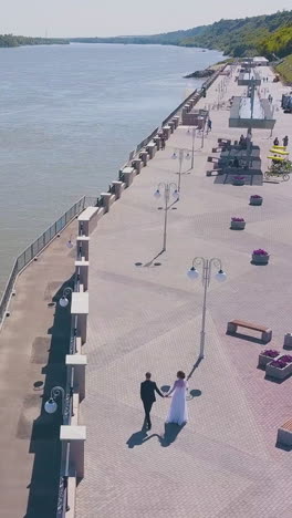 distant just married couple walks along modern waterfront of wide calm river on sunny day aerial view