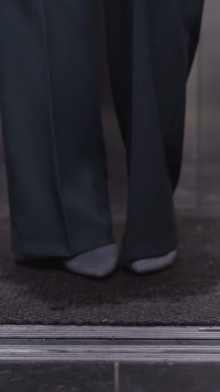 Person stepping out of elevator wearing business attire, focus on legs and shoes, close-up shot showing steps in professional setting, office, movement in modern building interior
