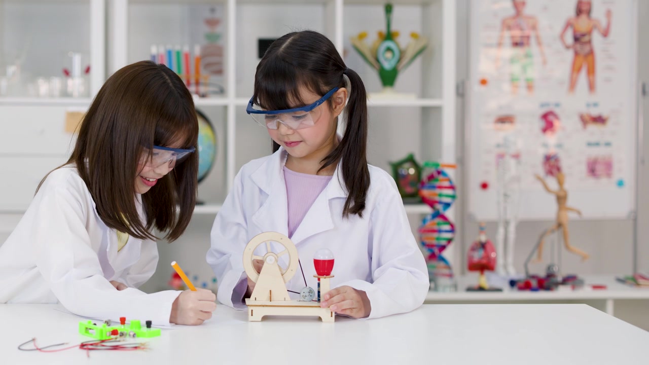 Two girls wearing safety goggles and lab coats study engineering toys in a bright classroom.