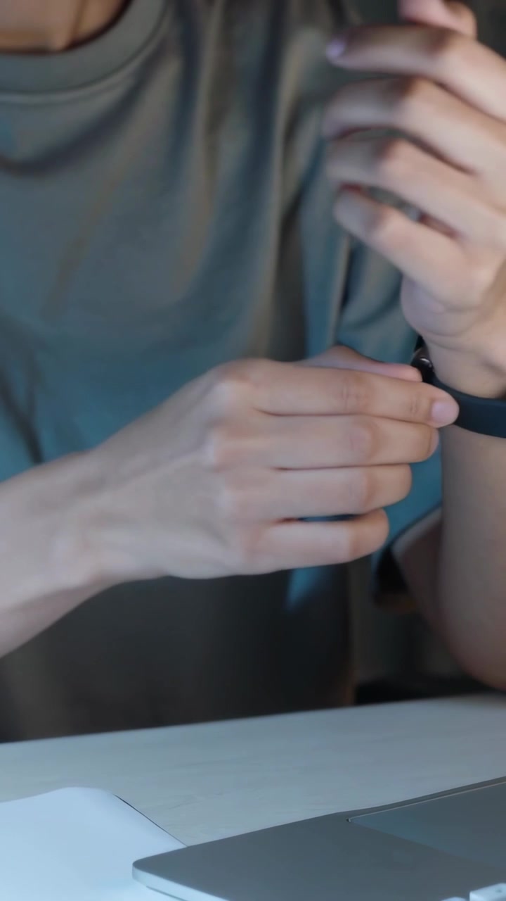 Video verticale: Donna con una maglietta grigia che guarda lo schermo, sistema l'orologio alla scrivania, con laptop e documenti, mentre si prepara.