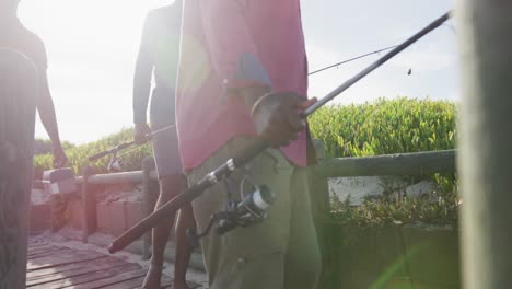 padre mayor afroamericano y dos hijos adolescentes con cañas de pescar caminando por la playa