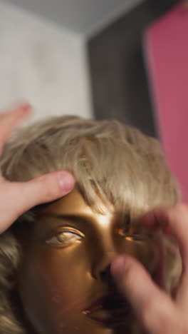 pov de una persona ajustando el peinado de una cabeza de maniquí dorada, dándole un toque de apoyo, con un fondo borroso