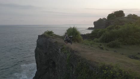 Aerial-orbits-male-motorcycle-tourist-atop-tall-ocean-cliffs-in-Bali