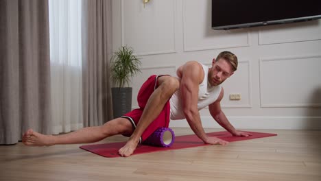 A-confident-blond-man-of-athletic-build-with-a-beard-in-a-white-T-shirt-and-red-pants-massages-his-thigh-during-morning-exercises-using-a-massage-roller-on-the-Red-carpet-in-a-modern-bright-apartment-at-home