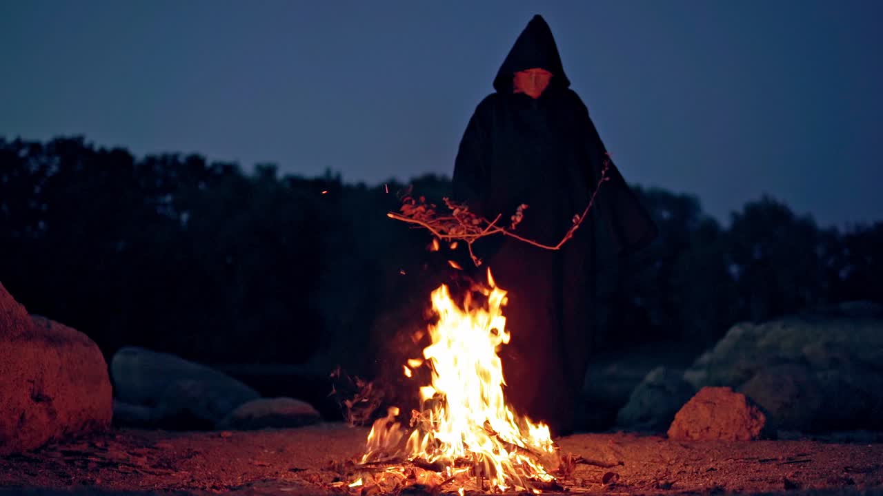 Witch makes scary ritual in the fire. Mysterious wizard in black cloak doing shamanism over the bright fire flame in nature at night.