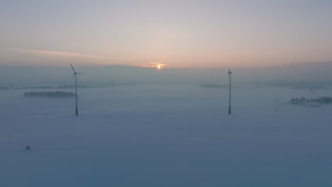 Luftaufnahme-Von-Windkraftanlagen,-Die-Erneuerbare-Energie-Im-Windpark-Erzeugen,-Schneebedeckte-Landschaft-Mit-Nebel,-Sonniger-Winterabend-Mit-Goldenem-Stundenlicht,-Breite-Drohnenaufnahme,-Die-Sich-Rückwärts-Bewegt