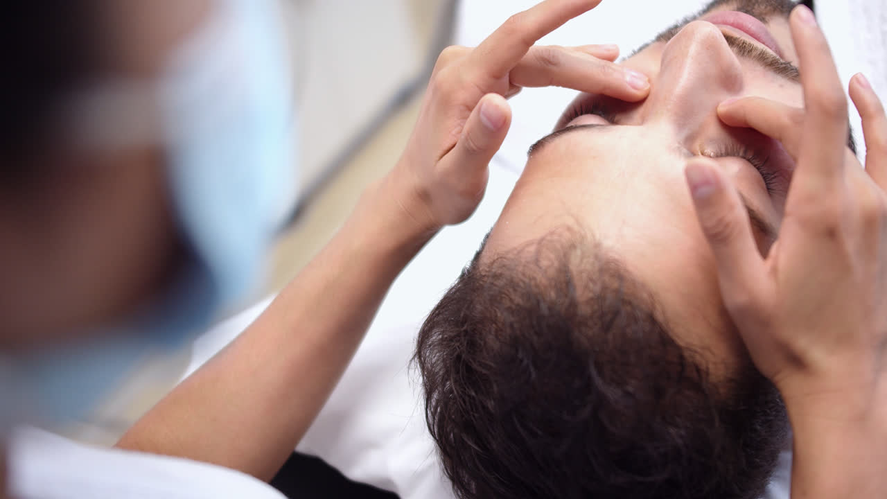 Beautiful slow motion close-up shot of a young man's face getting his dark circles massaged before receiving his beauty treatment lying on a massage table in a dermatology clinic.