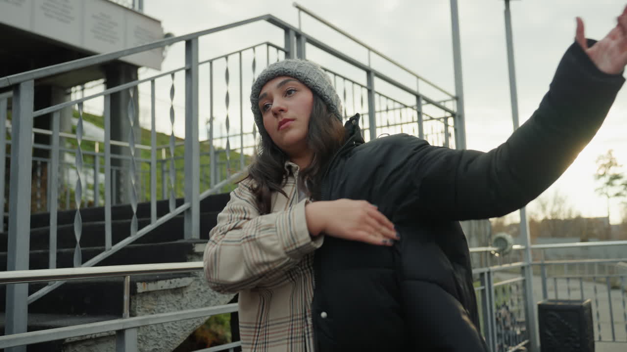 Donna che indossa un piumino vicino a scale metalliche al tramonto, preparandosi al riscaldamento per la street dance, sistema il berretto di lana, movimenti espressivi delle braccia, camicia a quadri a strati, controluce suggestivo, atmosfera di prova spontanea.
