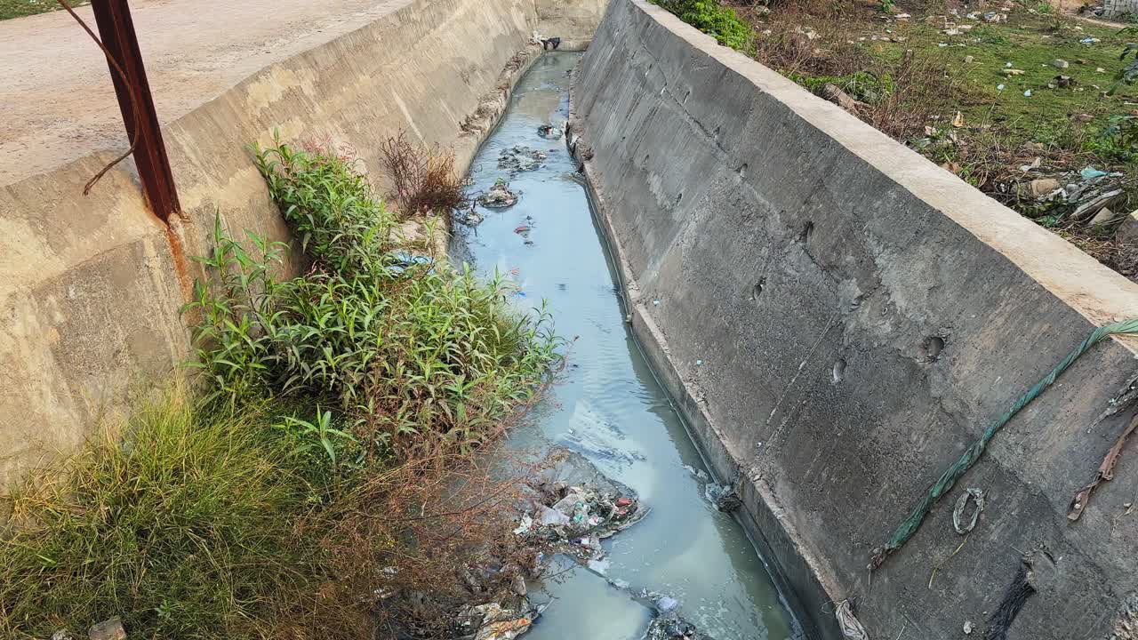 Plano de seguimiento de un canal de drenaje de hormigón que transporta aguas residuales turbias con residuos flotantes, desechos plásticos, proliferación de algas y maleza a lo largo de los bordes, mostrando la contaminación y el abandono urbano.