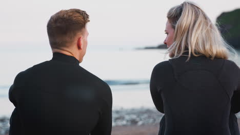 rear view of couple wearing wetsuits sitting on beach talking and looking out to sea together