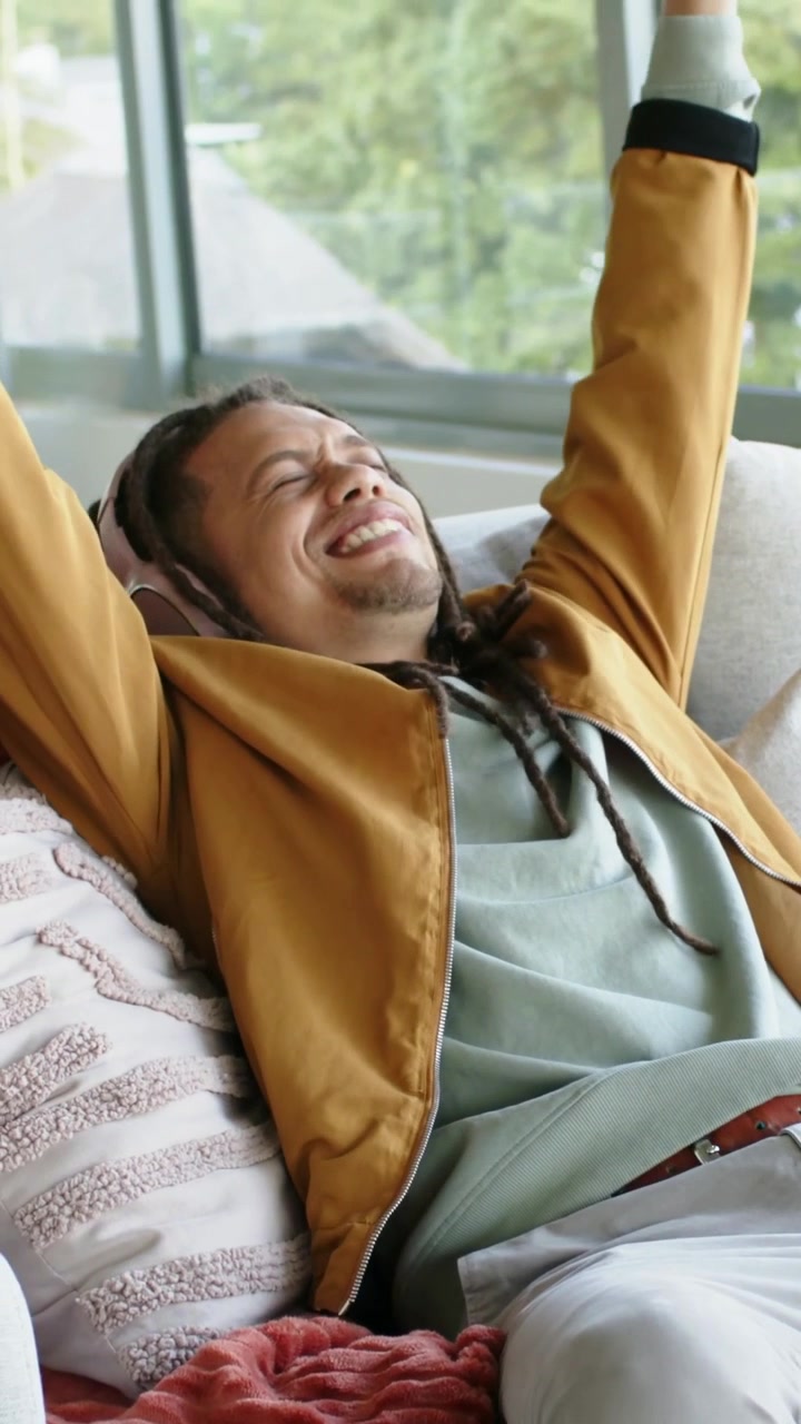 Video verticale: Musica in sottofondo, uomo con giacca dorata e cuffie che si allunga e si rilassa sul divano vicino alla finestra.