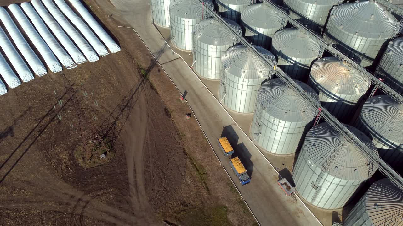 트럭이 채워진 노란 곡물은 새로운 금속 근처를 운전합니다. 은 날에 현대적인 곡물 엘리베이터. 공중 드론 뷰. 곡물 저장에 대한 큰 금속 둥근 실로 위의 수확 및 저장 비행 뷰. 큰 곡물 창고.