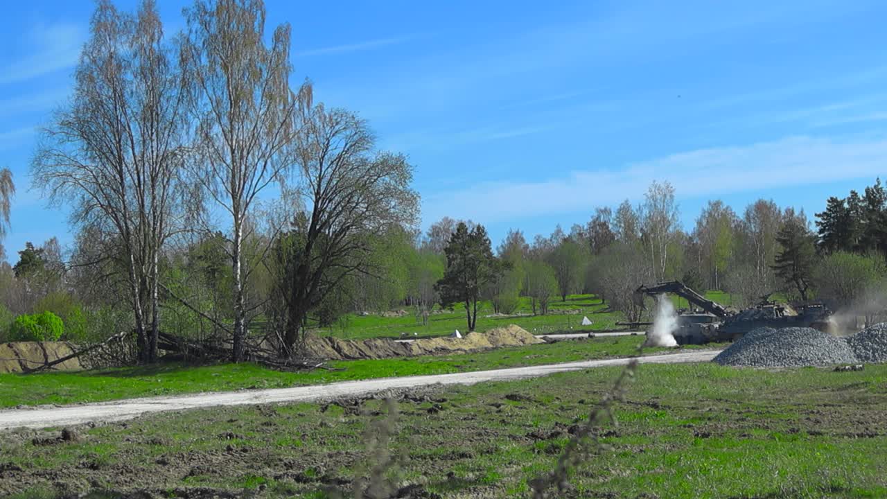 Rare slow motion footage of two British Challenger 2 4034 tanks on a sunny green battlefield. One is aming into the distace while the other moves with an excavator arm and approaches trenches.