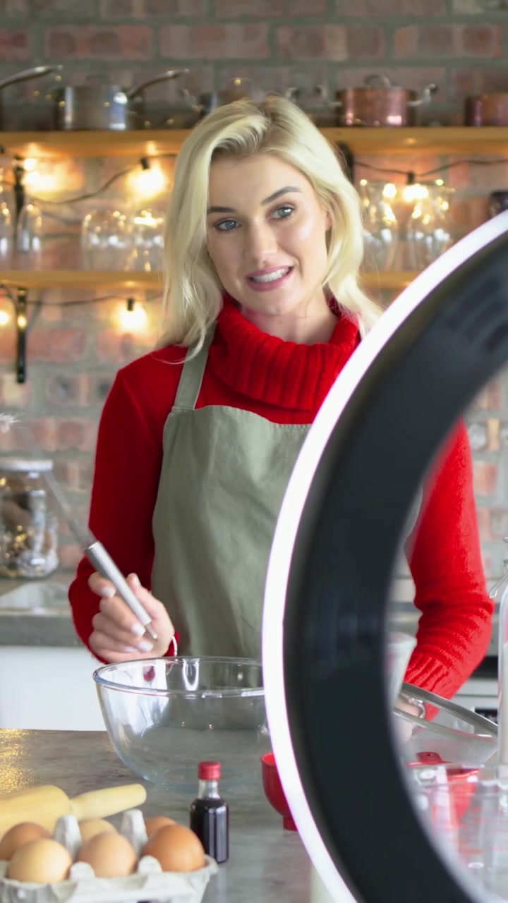 Video verticale: Donna che sorride alla telecamera, con top rosso e grembiule verde oliva, che sbatte una ciotola in cucina, mentre insegna.