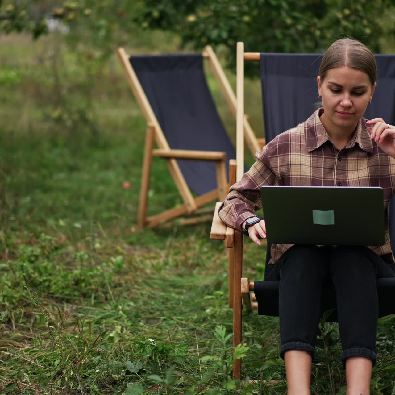 Pensive lady in plaid shirt sitting in garden chair working on laptop. Freelance working woman thinking over some work issues. Nature backdrop
