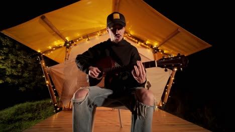 man plays the guitar and sings songs in a tent with light bulbs