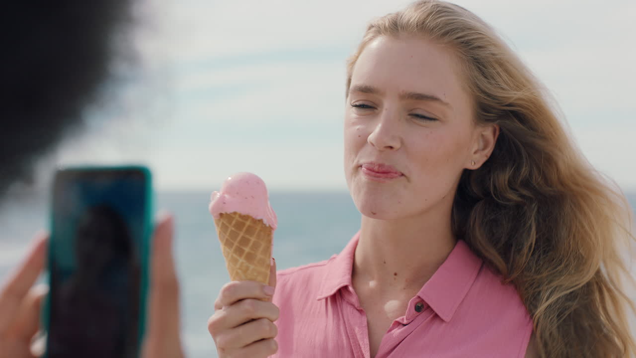hermosa mujer rubia con afro comiendo helado en la playa posando para una amiga tomando fotos usando un teléfono inteligente amigas compartiendo un divertido día de verano en las redes sociales 4k