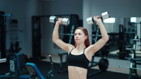 a young woman holds in her hands iron dumbbells and raises it on the background of sports equipment. pumping the biceps in the gym.