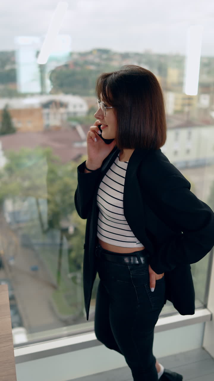 Slim attractive Caucasian woman with dark hair stands at the window. Lady speaks on the phone looking at city view. Vertical video.