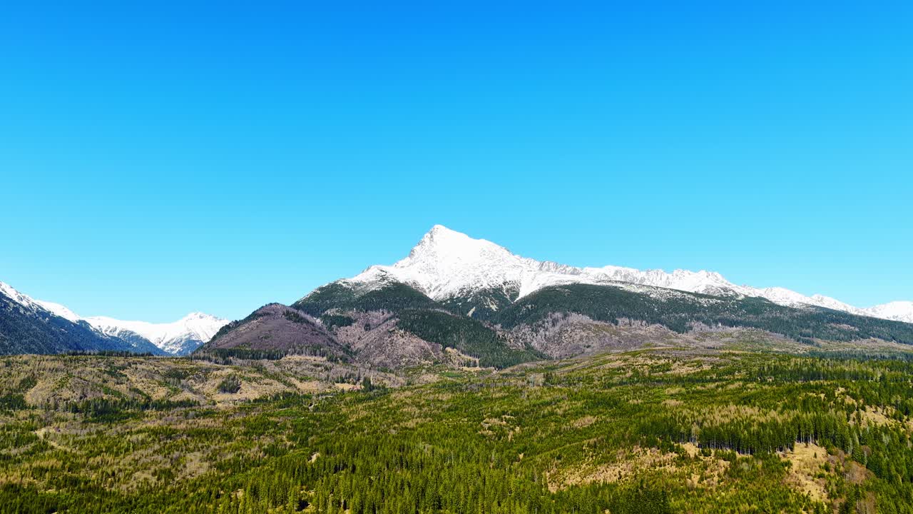 Lenta ripresa aerea cinematografica dell'iconica montagna Kriváň negli Alti Tatra, Slovacchia. Ampio paesaggio con foresta e colline che conducono a una cima innevata sotto un cielo azzurro limpido, ideale per riprese di viaggio e natura