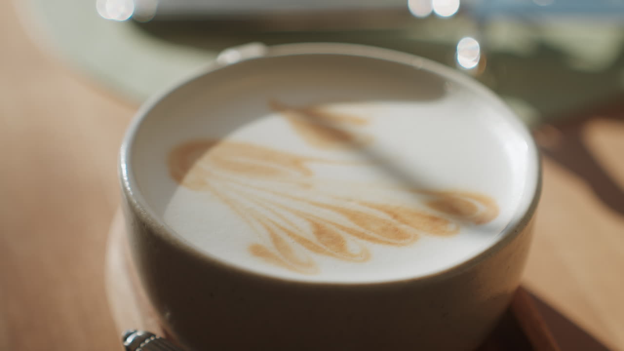 Tazza di latte illuminata dal sole con latte art, tazza in ceramica su un vassoio di legno, luce soffusa dalla finestra che proietta un'ombra delicata sulla schiuma di latte setosa e sul disegno al caramello, atmosfera mattutina accogliente e minimale, profondità di campo ridotta