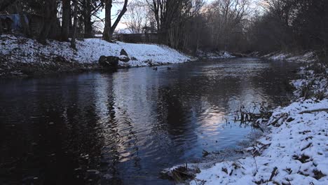 low angle footage of a dark brown river water slowly flowing and moving while camera is rising up and revealing more of the winter landscape surrounding the river. water is reflective, has ripples.