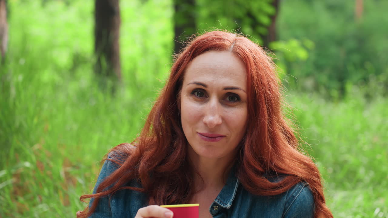 붉은 머리의 세련된 여성이 초록 숲, 햇살, 그리고 부드러운 바람으로 둘러싸여 평화로운 에너지가 가득한 한적한 피크닉 분위기를 즐기며 차분하게 휴식을 취하면서 야외에서 따뜻한 차를 마시고 있어요.