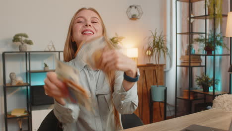 excited businesswoman freelancer holding fan of euro cash and dancing sitting at home office table
