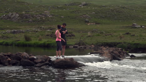 Pareja-Parada-Sobre-Rocas-En-Medio-De-Un-Río-Que-Fluye