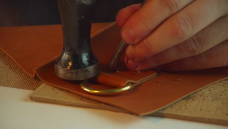 close-up of the tailor's hands punching holes for thread in the leather loop of the bag handle with a hammer and awl, slowmotion