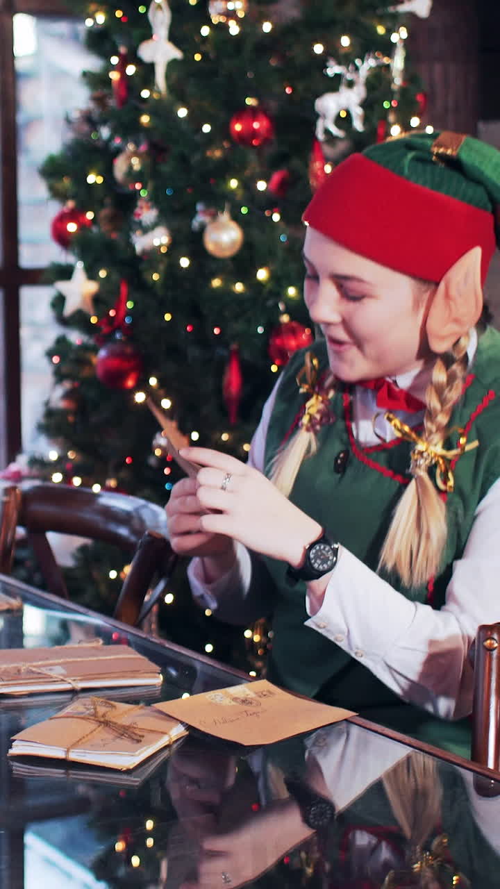 An elf girl sits in a room with a Christmas tree and looks through the letters to the children of Santa Claus. Vertical video