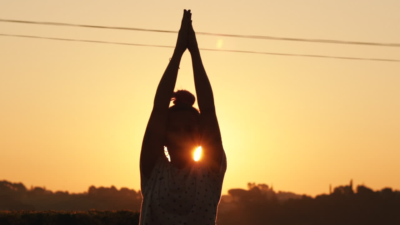 Steady shot of the silhouette of young woman in a sun salutation yoga pose at sunrise with a strong sun backlight