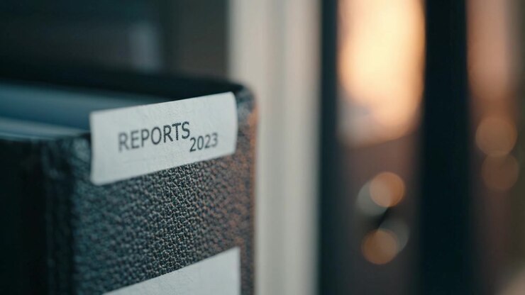 Primo piano di un raccoglitore di documenti etichettato 'Rapporti 2023' su uno sfondo sfocato, che evidenzia l'organizzazione dell'ufficio e la gestione degli archivi.