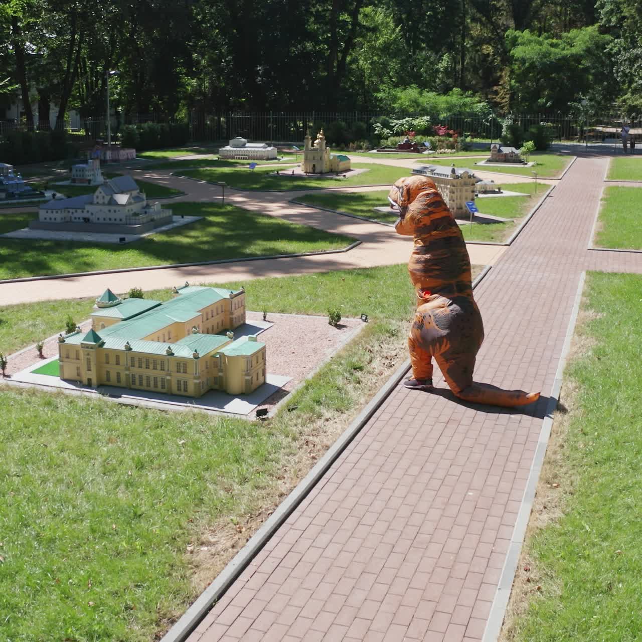 Person in air costume of dinosaur walking along the miniature buildings. Tirex creature having a walk between the outdoor museum on sunny summer day