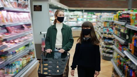 십대 소녀와 그녀의 어머니 또는 여동생이 마스크를 착용하여 카트와 함께 슈퍼마켓에서 쇼핑하고 있습니다.