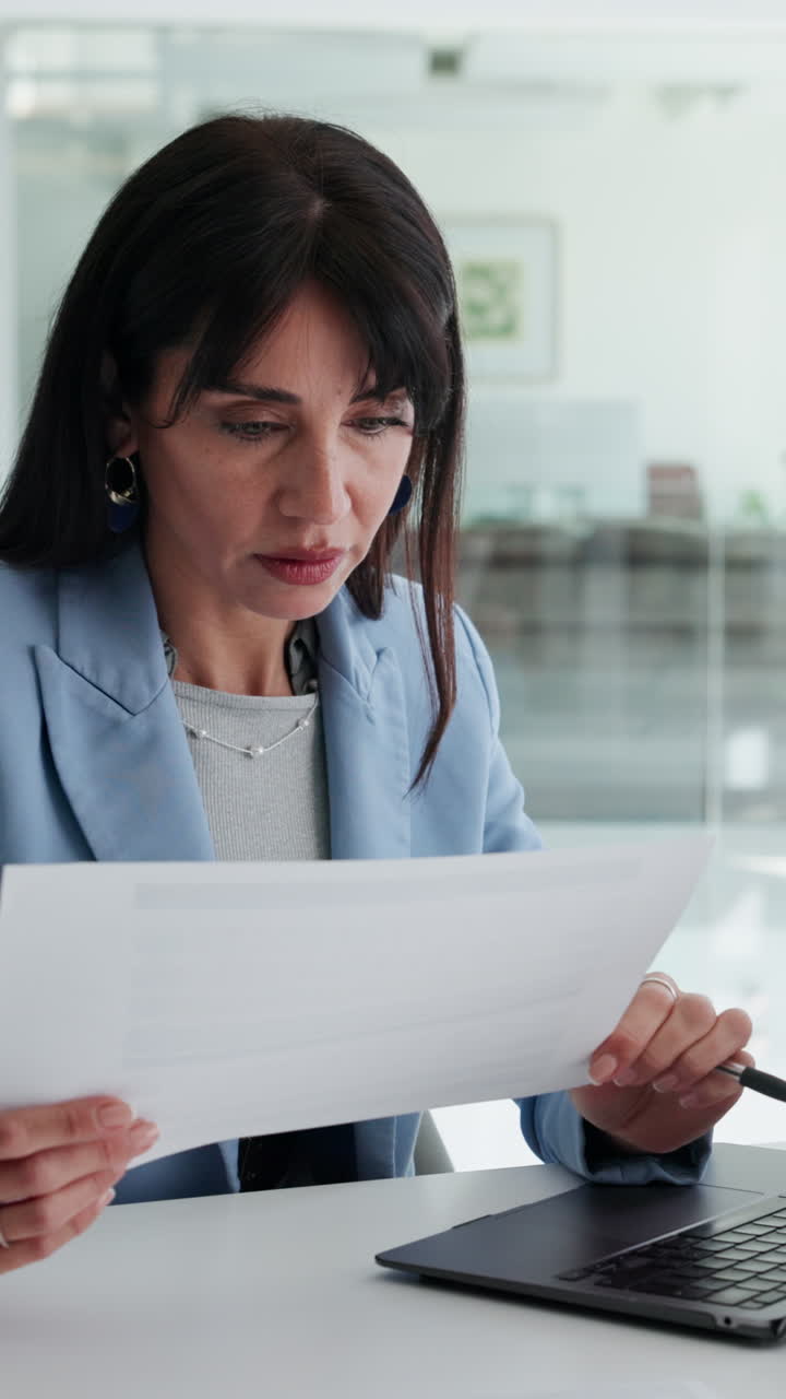 Mujer profesional leyendo un documento y escribiendo en su ordenador portátil en la oficina.