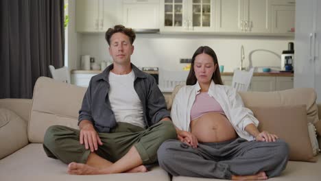 a happy man in a gray shirt together with his pregnant brunette wife sit on the sofa in the lotus position and meditate and then look at each other during their quiet time and waiting for the child at home