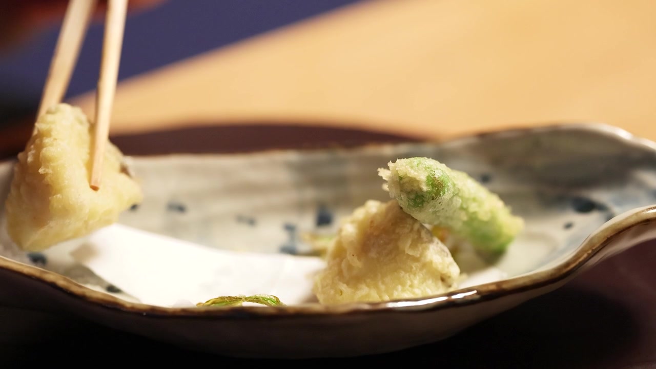 木製の箸が、伝統的な陶器の皿からサクサクの野菜天ぷらを持ち上げるクローズアップショット。