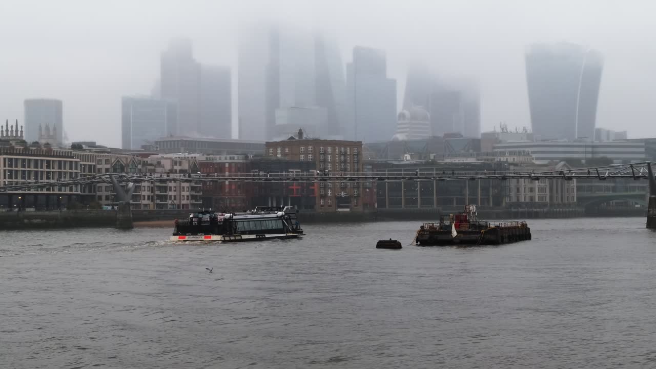 Uber Boats sail under the Millennium Bridge on the River Thames, with the City of London financial district skyscrapers in the background during a misty rainy day in UK’s vibrant capital city.