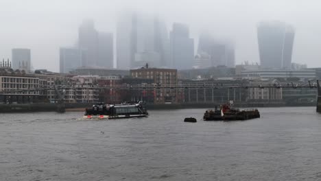 uber boats sail under the millennium bridge on the river thames, with the city of london financial district skyscrapers in the background during a misty rainy day in uk’s vibrant capital city.