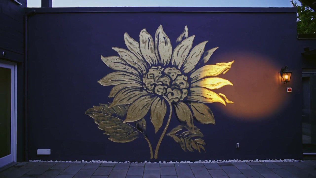 La telecamera si allontana rivelando un murale di girasoli dorati su una parete in un cortile, mostrando un riflettore che si affievolisce.