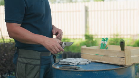 close up farmer at yard drops work tool onto blue drum beside wooden crate with gloves, apron belt on waist visible, summer daylight scene near house fence