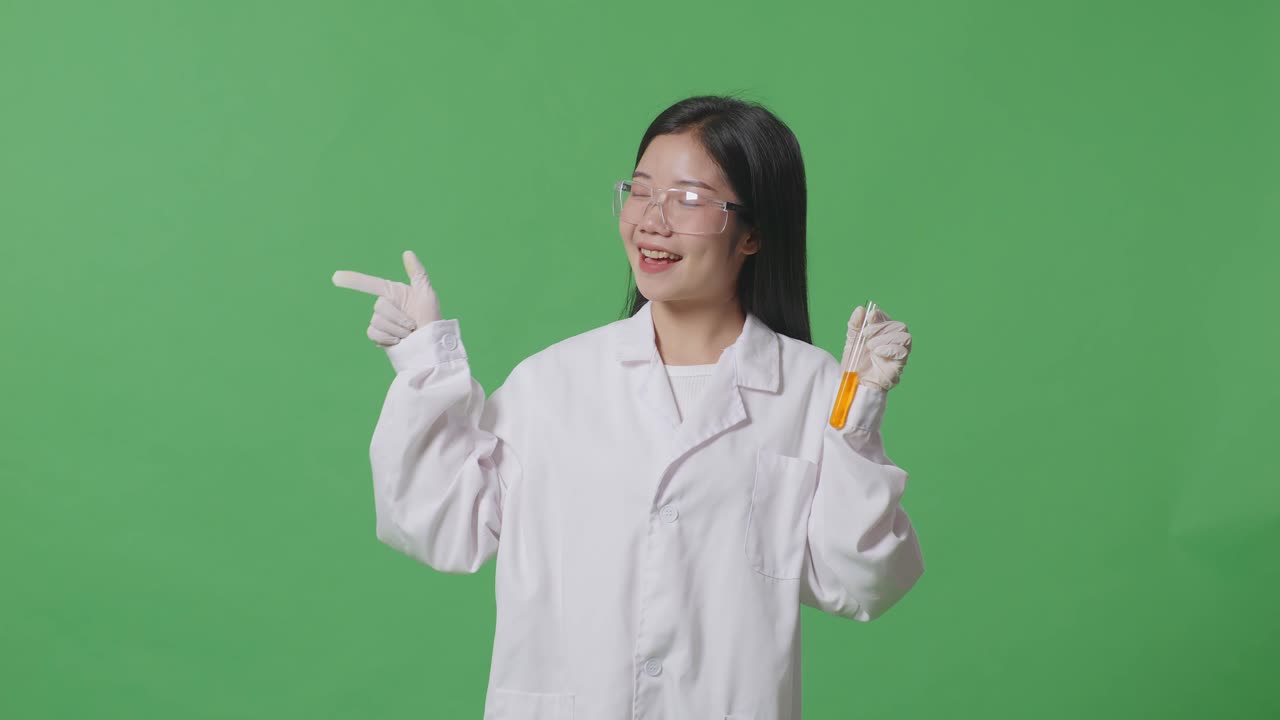 실험실에서 녹색 화면 배경에 서 있는 동안 미소 짓고 으로 가리키는 시험관에서 오렌지 액체와 함께 아시아 여성 과학자