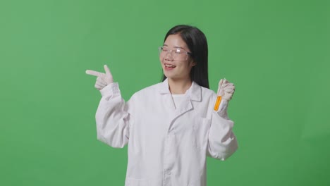 asian woman scientist with orange liquid in the test tube smiling and pointing to side while standing on the green screen background in the laboratory